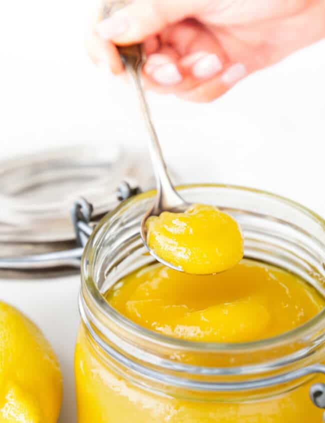 A hand holding a spoonful of lemon curd over a jar of lemon curd.