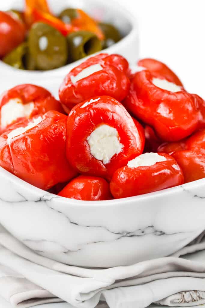 a white marble bowl of Stuffed Cherry Peppers and a cloth napkin under the bowl and a bowl of olives in the background.