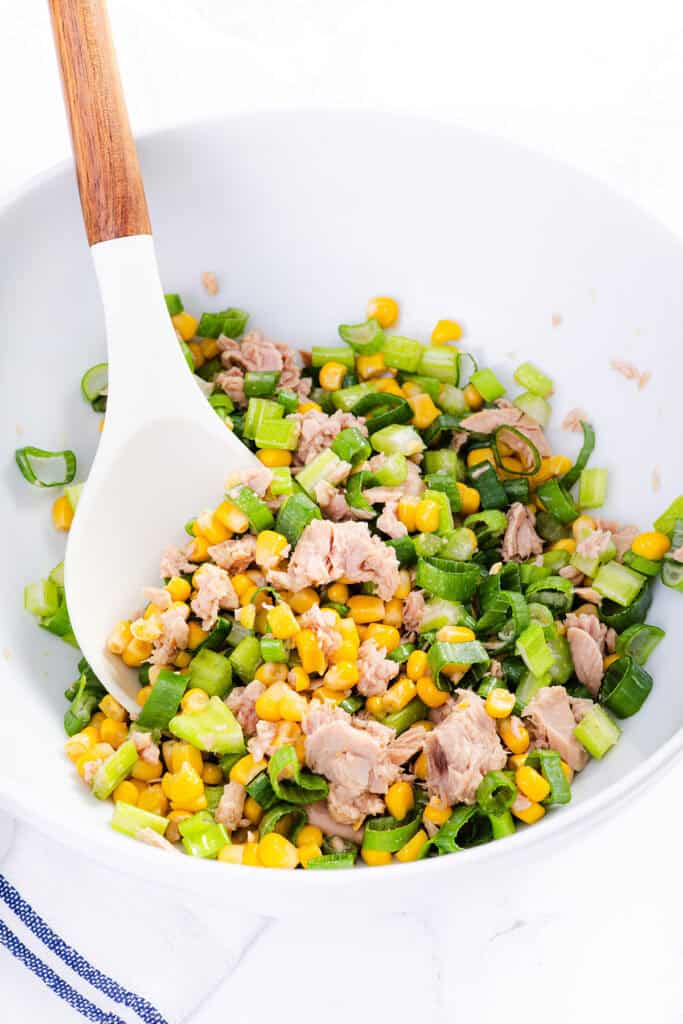 A white bowl contains a mixture of chopped green onions, corn, and chunks of tuna, resembling a fresh Tuna Pasta Salad, with a white spoon featuring a wooden handle resting inside.