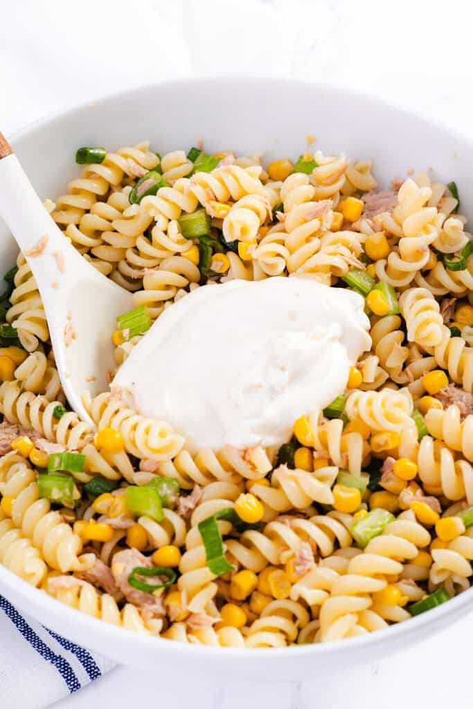 A white bowl filled with Tuna Pasta Salad—rotini pasta, corn, green onions, and tuna—topped with a dollop of creamy white sauce, with a spoon resting in the mixture.