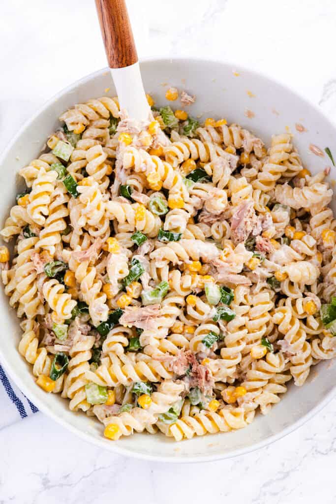 A bowl of Tuna Pasta Salad made with rotini, tuna, corn, green onions, and a creamy dressing. A spoon with a wooden handle rests in the bowl, ready for serving this flavorful dish.