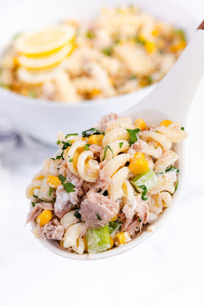 A close-up of a spoonful of creamy Tuna Pasta Salad with corn, chopped celery, and parsley, held above a bowl of the same salad, beautifully garnished with lemon slices.