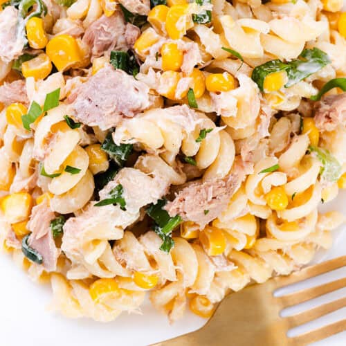 A close-up of a Tuna Pasta Salad featuring rotini mixed with corn, tuna, and chopped greens, with a gold fork placed on the side.