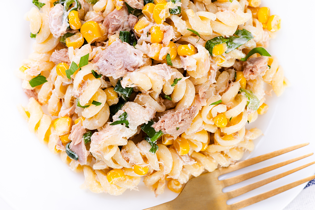 A close-up of a Tuna Pasta Salad featuring rotini mixed with corn, tuna, and chopped greens, with a gold fork placed on the side.