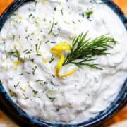 a close up image of a bowl of Tzatziki topped with chopped dill and lemon zest and surrounded by pita bread.