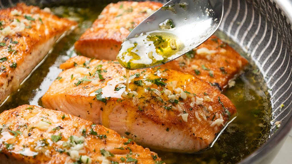 A metal spoon drizzles buttery garlic sauce over pan-seared salmon fillets in a skillet.