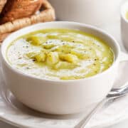 A bowl of creamy celery soup topped with celery pieces and black pepper, served on a white plate with a spoon, with a basket of sliced bread in the background.