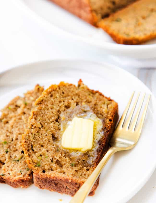 A slice of zucchini bread with a pat of melting butter on top, served on a white plate with a gold fork to the side.