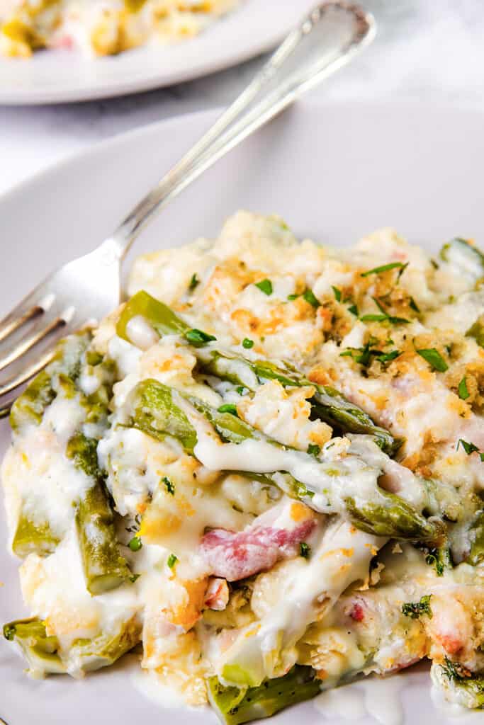 A plated serving of asparagus casserole on a white plate, with a fork digging in. The casserole is a blend of bright green asparagus pieces and chopped bacon in a creamy white sauce, topped with melted cheese and crispy golden breadcrumbs, garnished with chopped parsley.