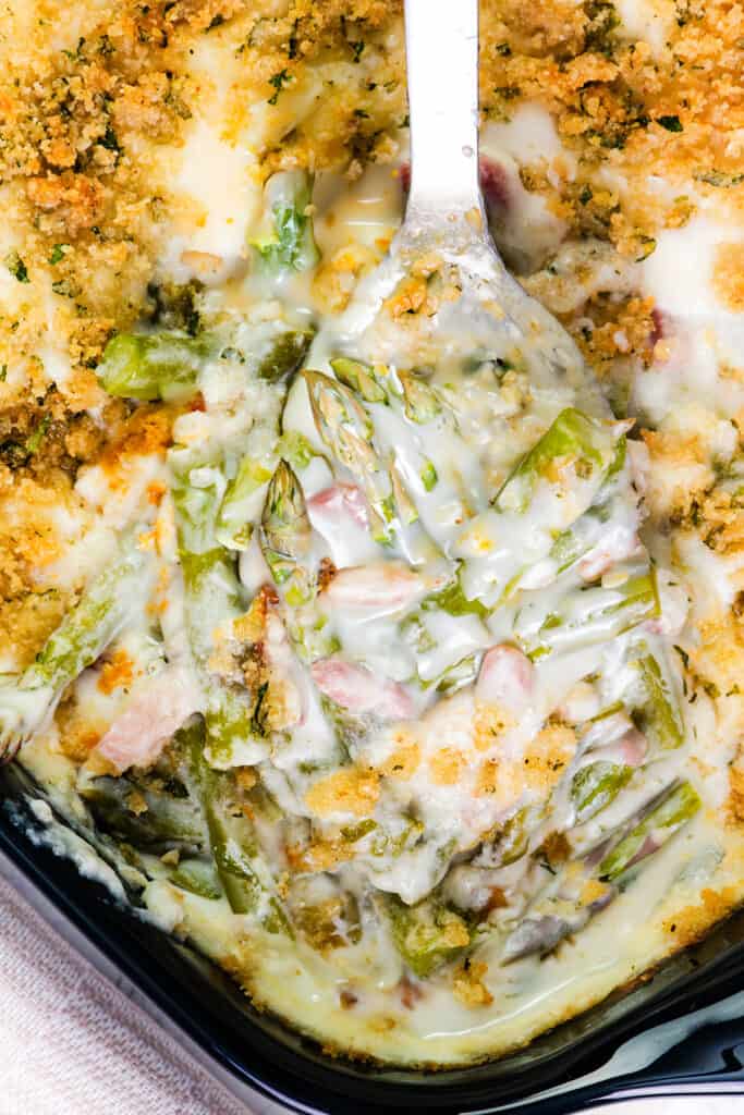 Close-up of a creamy asparagus casserole in a black dish, featuring green asparagus tips and sliced ham covered with melted cheese and a golden-brown breadcrumb crust, with a serving spoon lifting a portion to reveal the stringy cheese and tender layers.