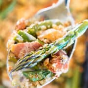 A spoonful of asparagus casserole held above a baking dish, with focus on the creamy mixture of chopped asparagus spears, bacon pieces, melted cheese, and golden breadcrumbs, all captured in sharp detail against a softly blurred background.