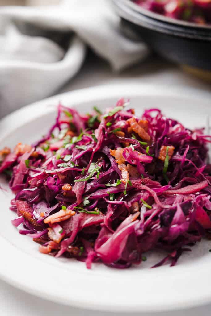 Braised purple cabbage topped with crispy bacon and garnished with chopped fresh herbs on a white plate.