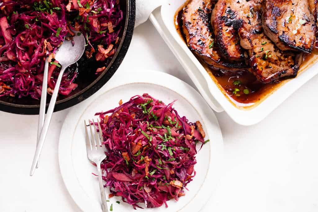 a plate of braised purple cabbage with a pan of cabbage and a platter of pork chops in the background.