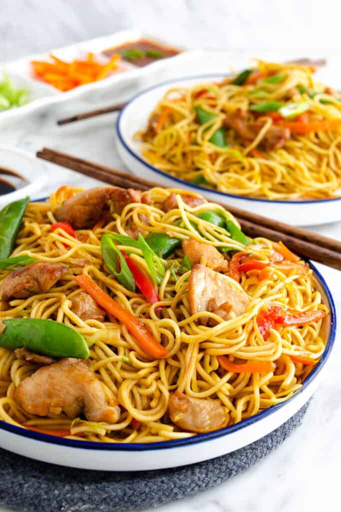 Two bowls of chicken lo mein with stir-fried noodles, pieces of chicken, bell peppers, carrots, and snow peas, with chopsticks.