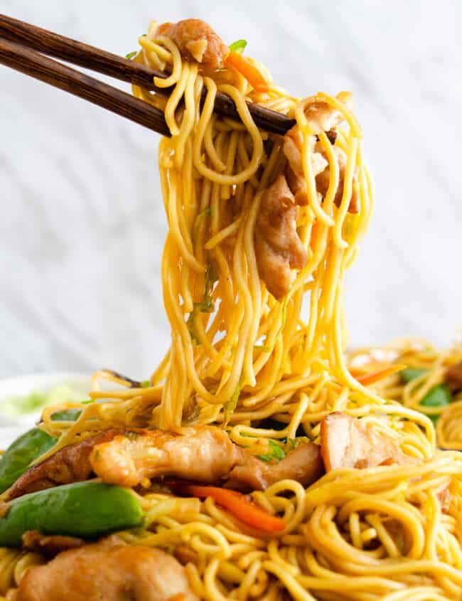 Chopsticks lifting strands of noodles, chunks of chicken, and mixed vegetables including carrots and green snap peas.