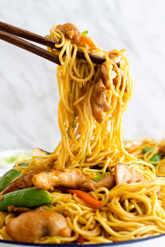 Chopsticks lifting strands of noodles, chunks of chicken, and mixed vegetables including carrots and green snap peas.