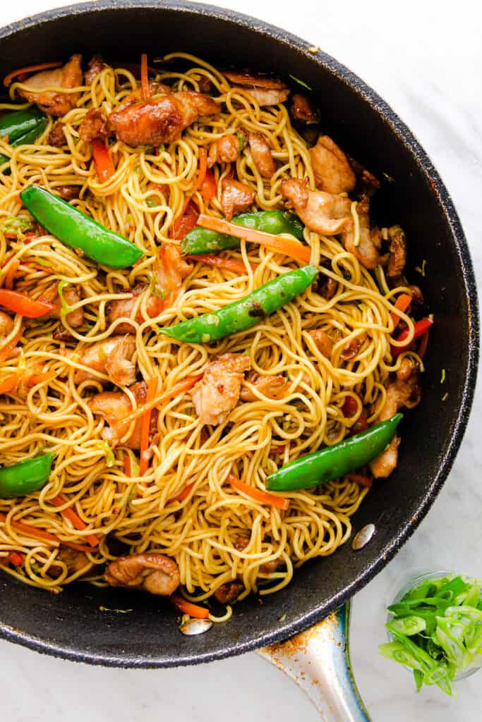 Chicken lo mein in a skillet with noodles, vegetables, and snap peas, garnished with green onions, on a marble surface.