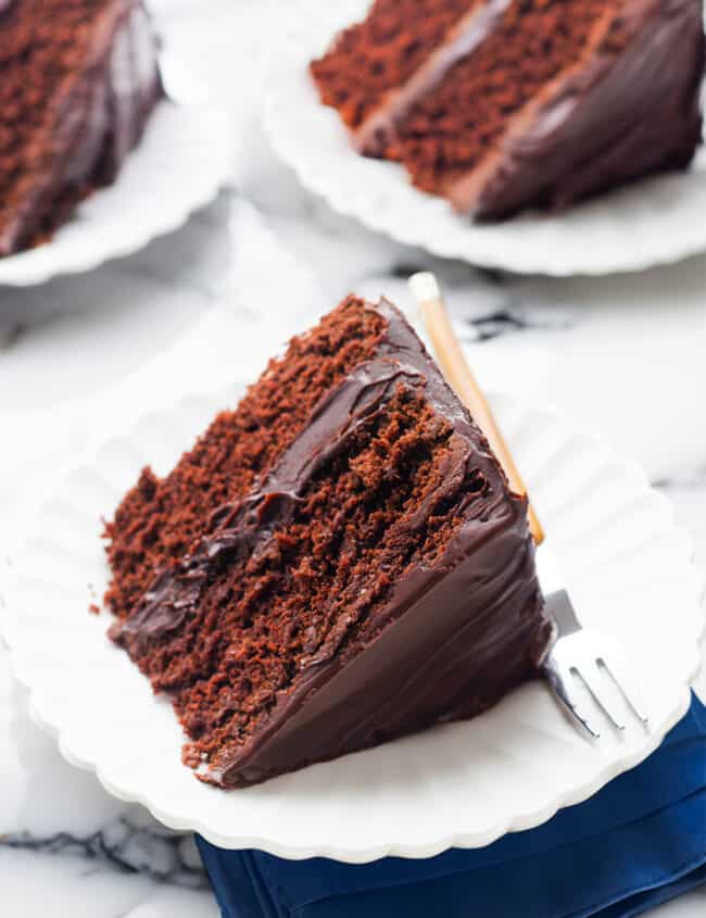 A slice of devil's food cake with glossy chocolate frosting on a white scalloped plate, with a fork on the side and a dark blue napkin underneath, set on a marble surface.