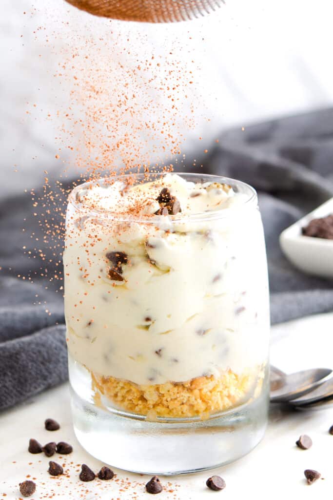 Cinnamon being sprinkled over a glass of ricotta dessert layered with graham cracker and chocolate chips, 