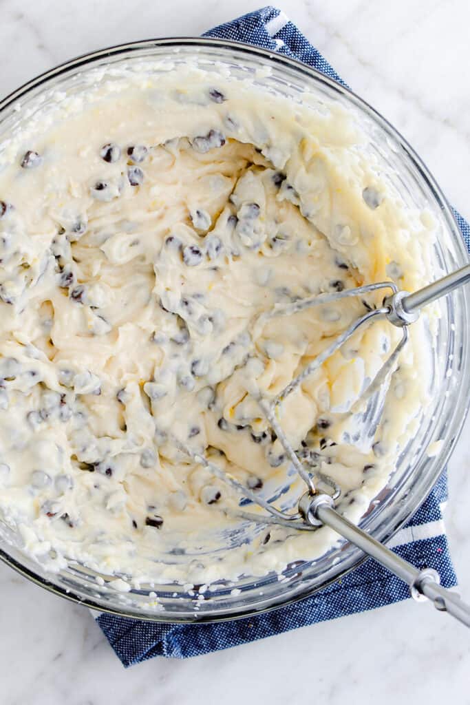 A bowl of rich ricotta cheese dessert filling speckled with chocolate chips, being mixed with a hand mixer, on a kitchen counter with a blue and white cloth underneath.