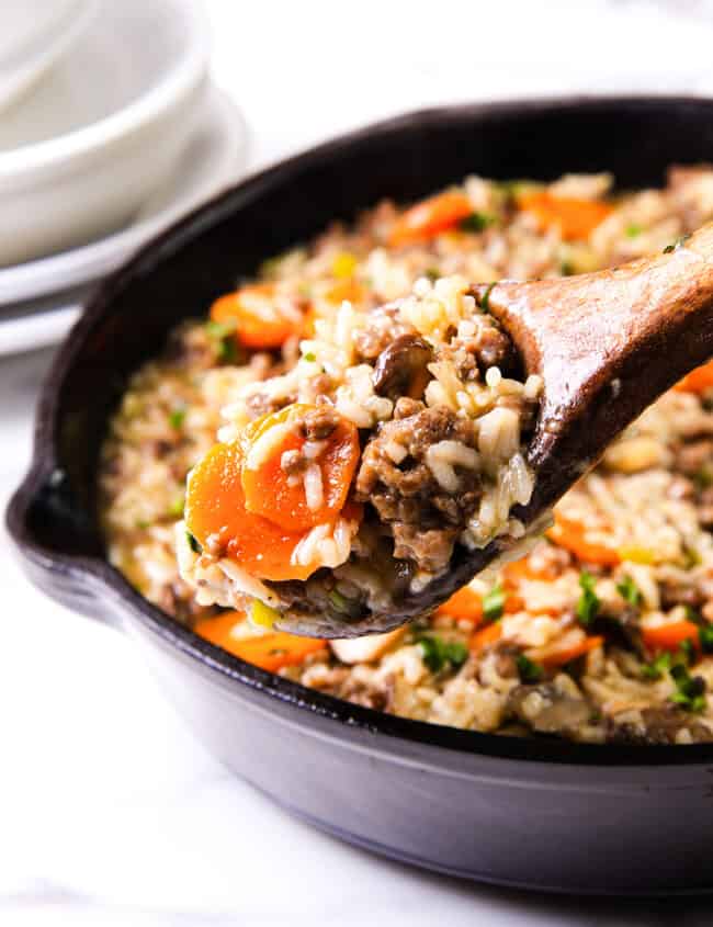 A wooden spoon scooping a portion of hamburger and rice with visible carrots and ground beef from a cast-iron pan.