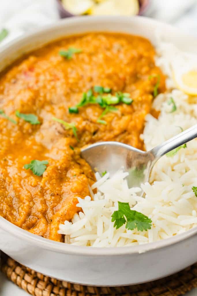 A close-up of a spoon resting in a bowl of Red Lentil Dahl curry garnished with fresh cilantro, next to a pile of white rice,