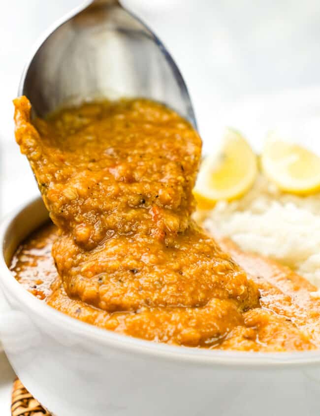 a close up image of a spoon serving up vibrant red lentil dal from a bowl, with fluffy white rice and lemon wedges in the background.