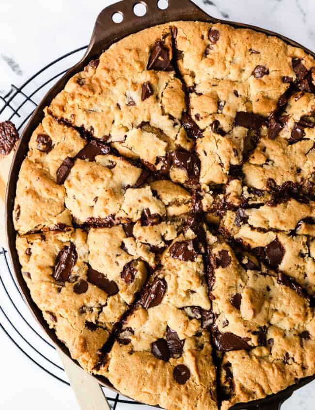 A freshly baked skillet cookie with gooey chocolate chunks, presented in a cast-iron pan on a wire cooling rack, with a wooden spatula.