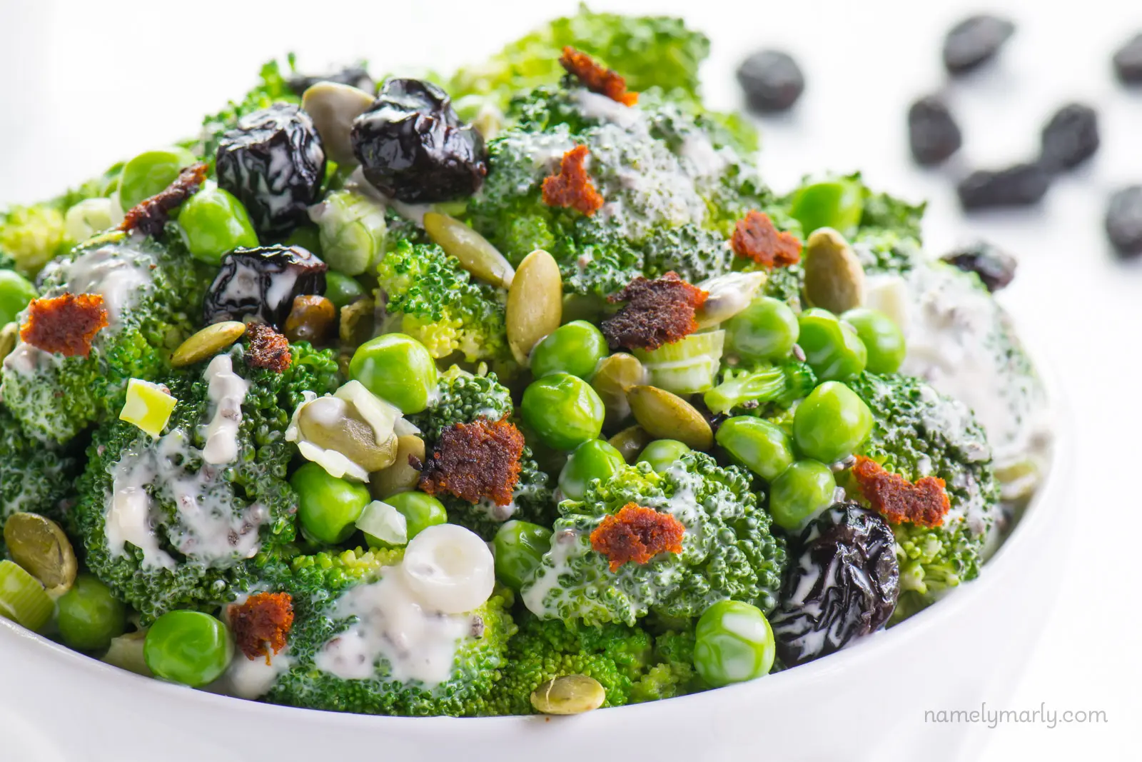 A white bowl filled with a broccoli salad made with broccoli florets, green onion, peas, and raisins.