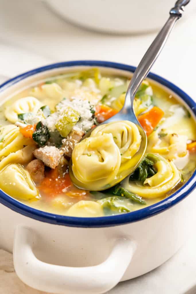 A close-up of a comforting Chicken Tortellini Soup in a white bowl with blue rim, with a spoon ready for enjoying.