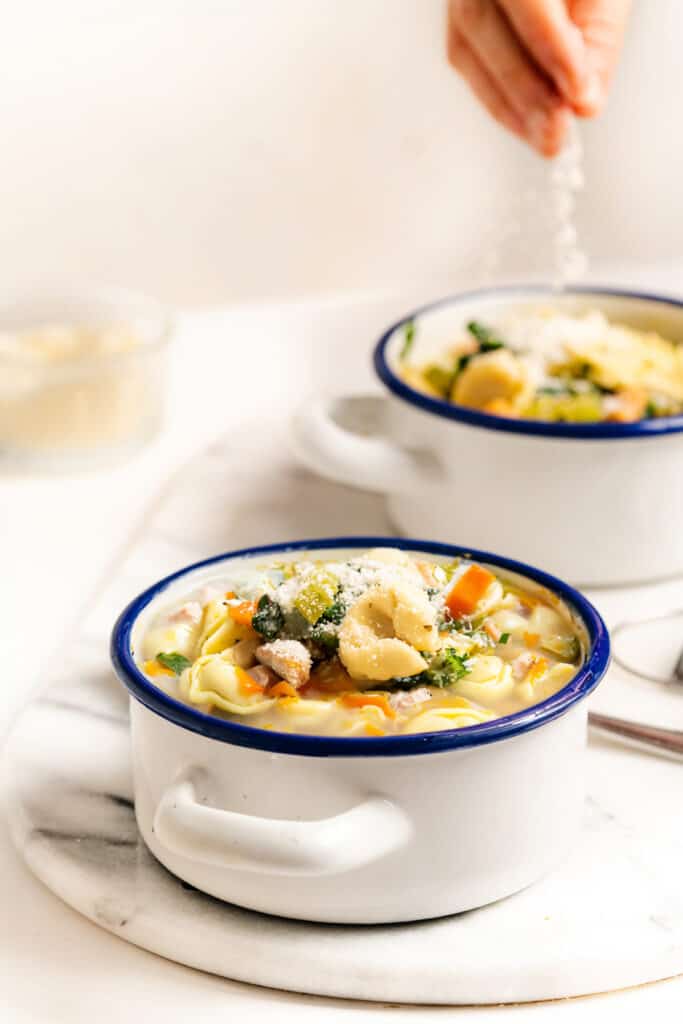 A hand sprinkling grated Parmesan cheese over a bowl of Chicken Tortellini Soup, filled with pasta, chicken, and vegetables.