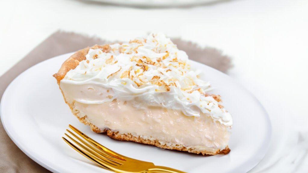 A slice of coconut cream pie on a plate with the pie in the background.