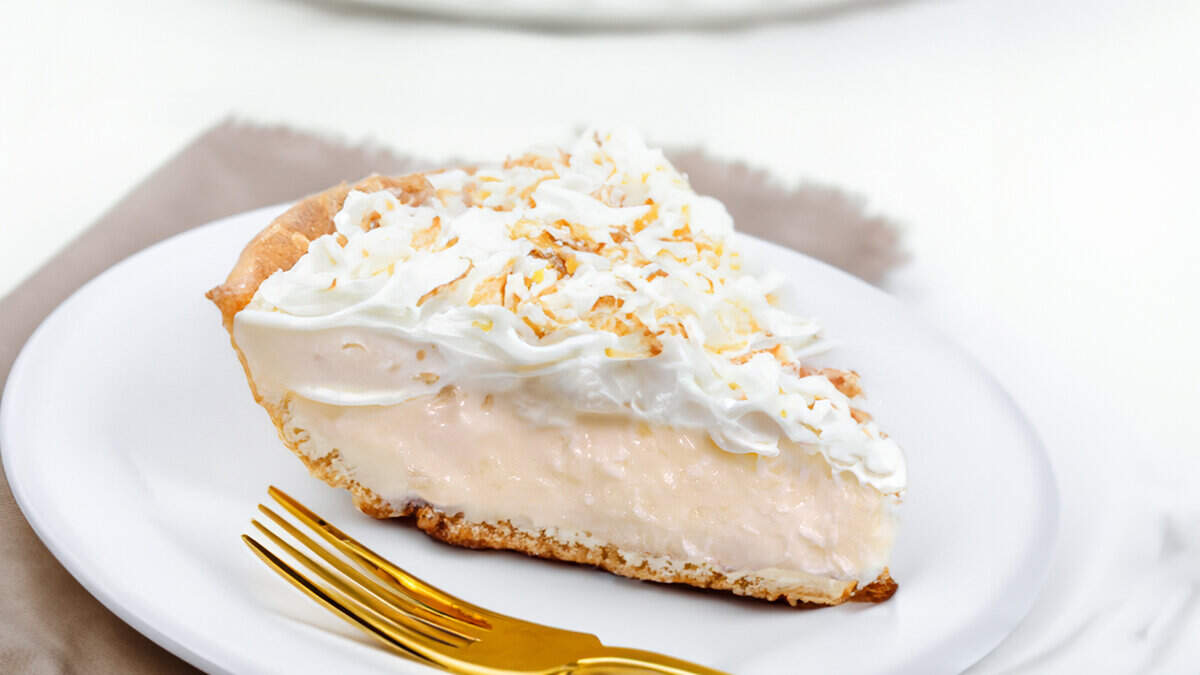 A slice of coconut cream pie on a plate with the pie in the background.