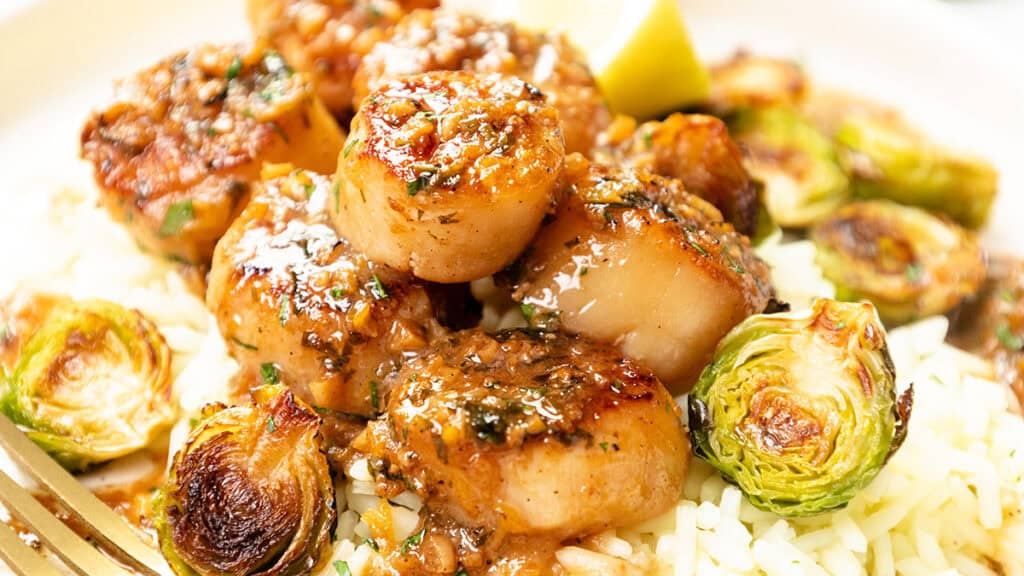 A close-up reveals seared scallops with a rich sauce atop rice, complemented by roasted Brussels sprouts. This delectable dish, from our "20 Easy Recipes" collection, is garnished with a lemon wedge for that perfect zest.
