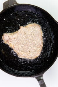 A breaded pork chop beginning to fry in oil in a cast-iron skillet.