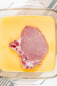 A raw pork chop soaking in an egg wash in a glass dish, prepping for breading.