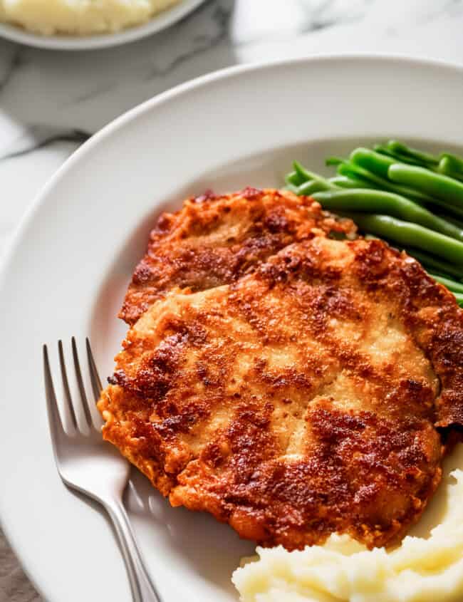 A crispy panko-breaded pork chop served with mashed potatoes and green beans on a white plate.