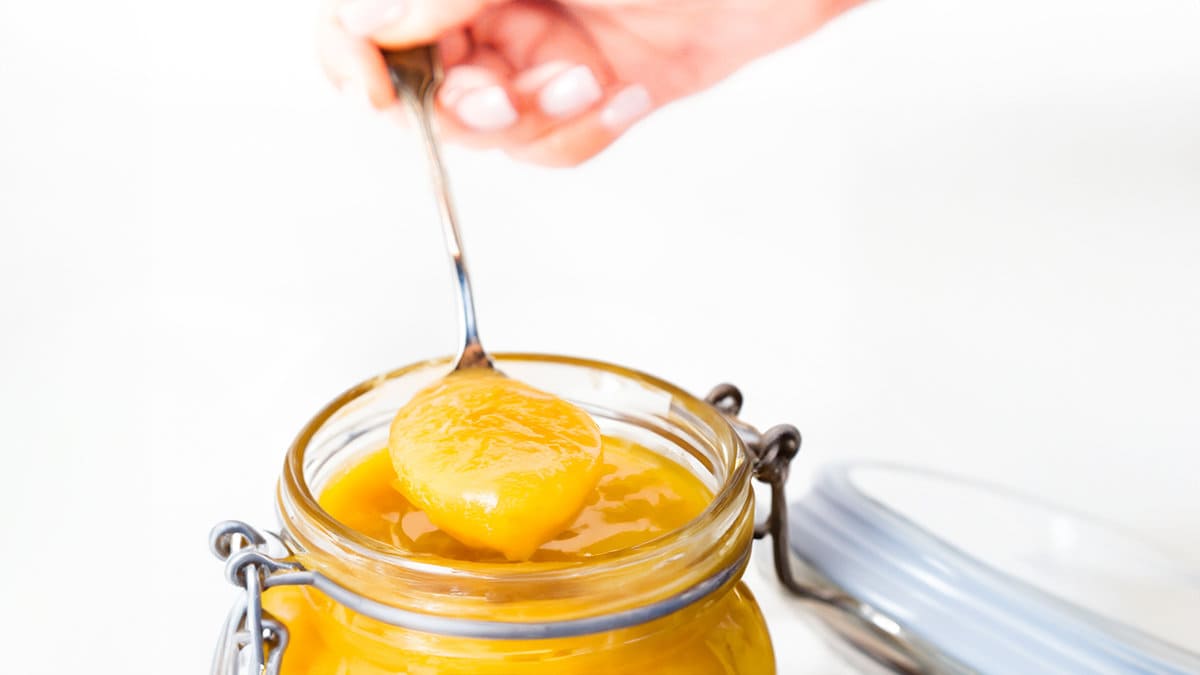 A hand holding a spoonful of lemon curd over a jar of lemon curd.