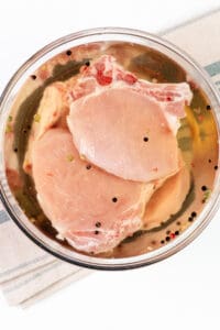 Pork chops marinating in a clear brine with peppercorns in a glass bowl, atop a striped kitchen towel.