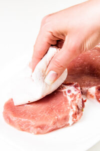 A hand with a pink manicure patting a raw pork chop dry with a paper towel on a white surface.