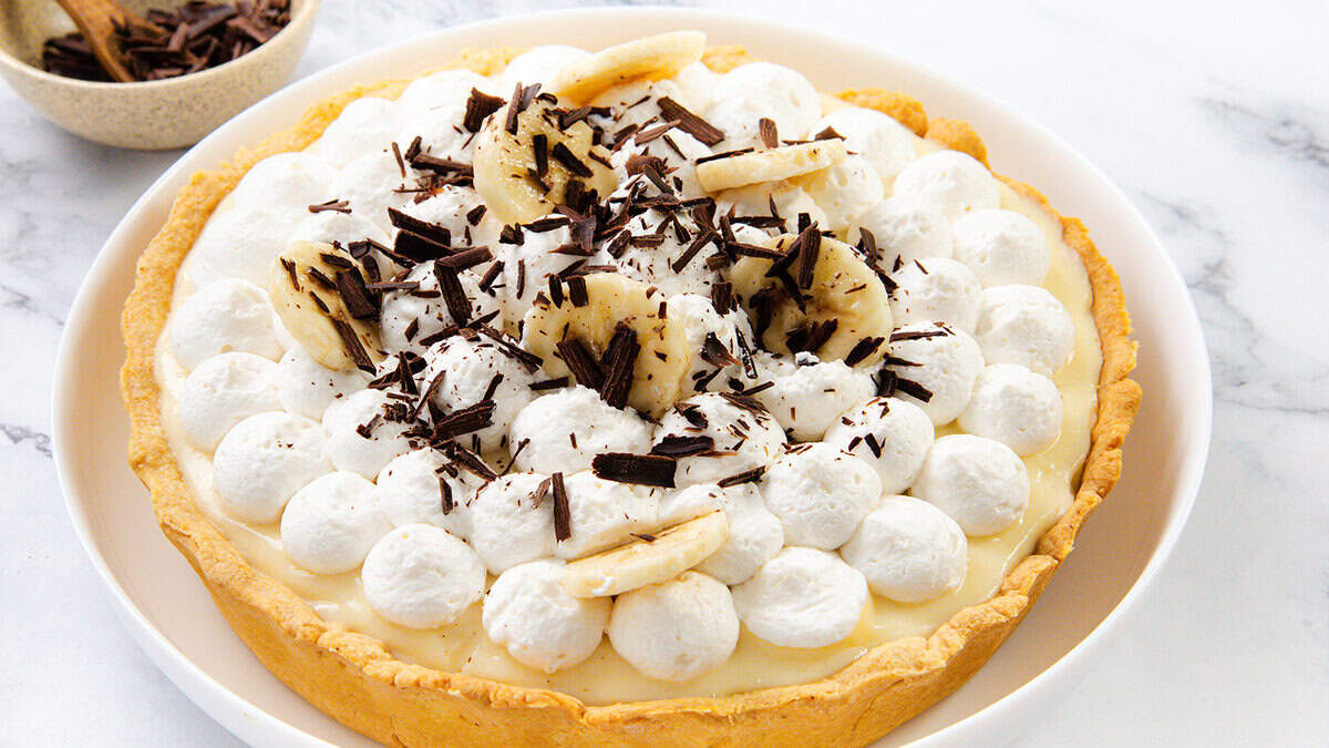 A banana cream pie topped with dollops of whipped cream, banana slices, and chocolate shavings, served on a white plate on a marble surface—a classic favorite among dessert recipes.