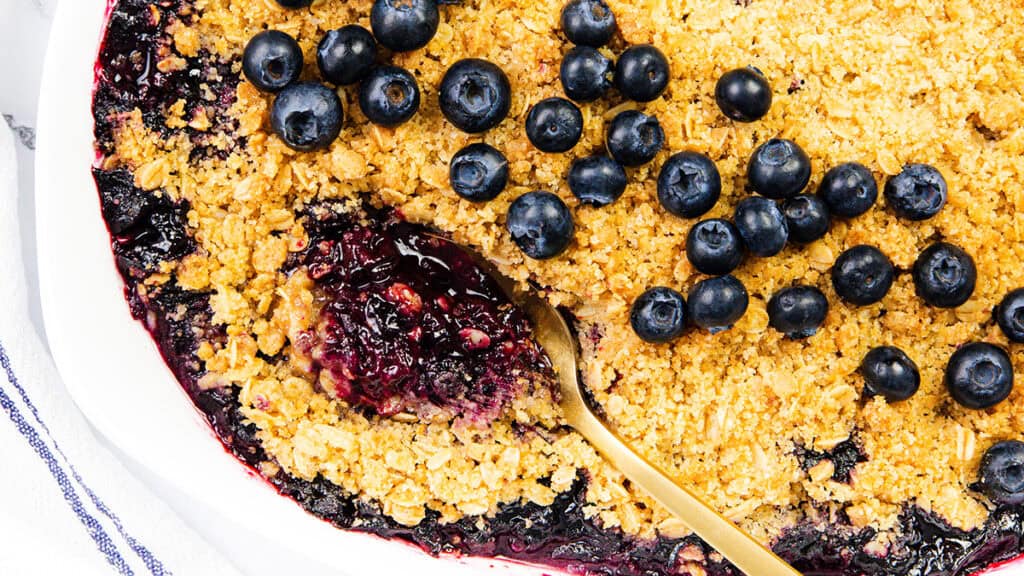 A close-up of a blueberry crumble topped with fresh blueberries and golden, crumbly oat topping. A gold spoon scoops out a portion, revealing the juicy blueberry filling—perfect inspiration for your Summer Dessert Recipes.
