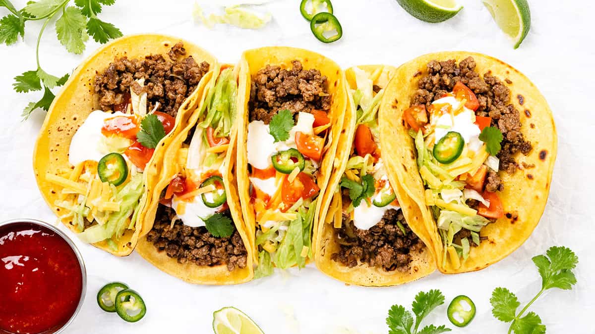 Four beef tacos in yellow corn tortillas topped with lettuce, shredded cheese, tomatoes, sour cream, sliced jalapeños, and cilantro—served with lime wedges and red salsa—make a perfect choice for Easy Dinner Recipes.