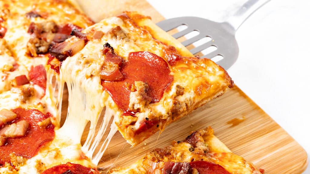 A slice of pepperoni and sausage pizza is being lifted from a wooden board, with melted cheese stretching from the slice to the rest of the pizza—a mouthwatering classic among Italian recipes.
