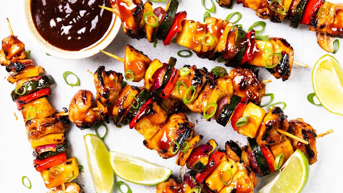 Grilled chicken and vegetable skewers glazed with sauce, garnished with chopped green onions, served with lime wedges and a bowl of dark dipping sauce on a white surface—perfect for Easy Dinner Recipes.