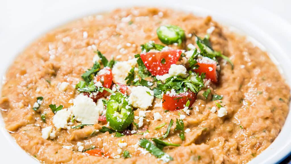 A bowl of refried beans topped with jalapenos, tomatoes, and crumbled cheese.
