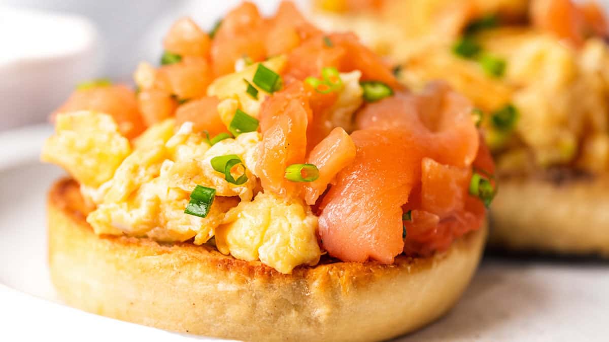 Two toasted English muffins topped with scrambled eggs and smoked salmon on a white plate.