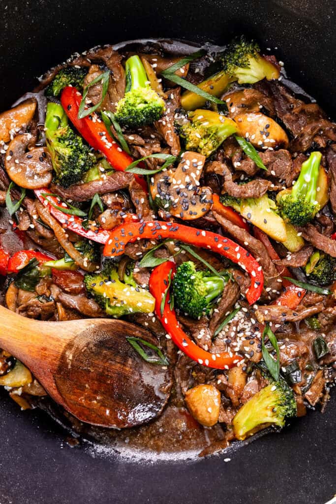 A sizzling steak stir fry with broccoli, mushrooms, and bell peppers, sprinkled with sesame seeds in a black skillet, accompanied by a wooden spoon.