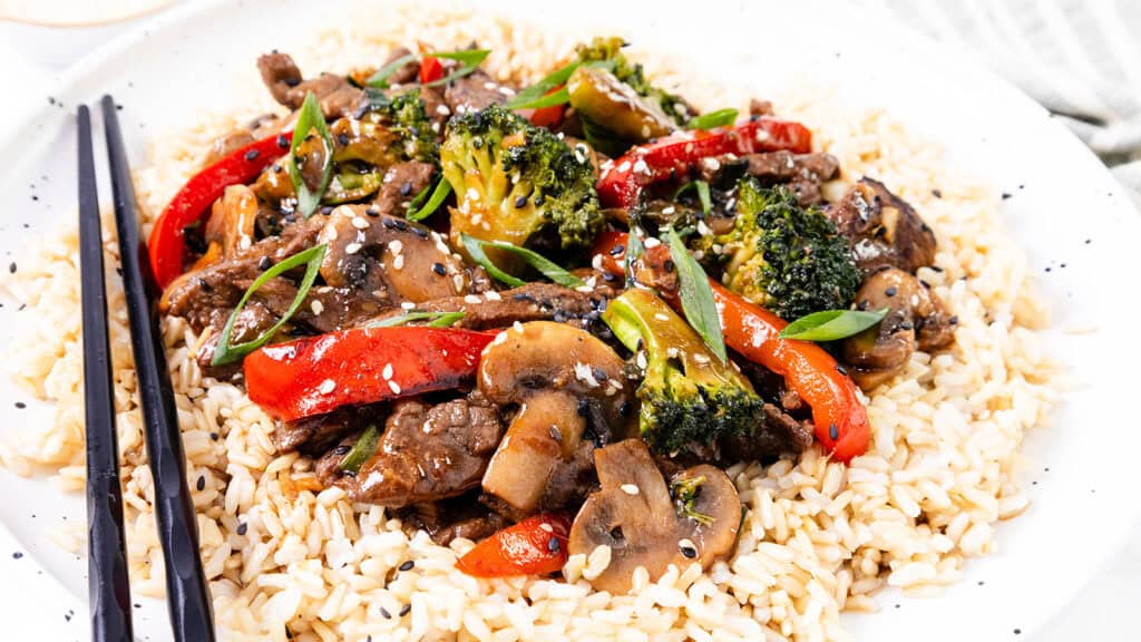 A steak stir fry with broccoli, red bell peppers, and mushrooms served over white rice, garnished with sesame seeds and accompanied by black chopsticks.