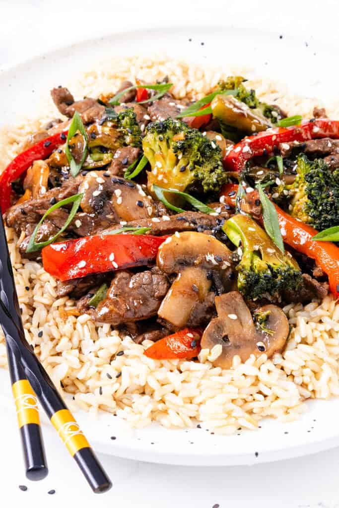 A steak stir fry with broccoli, red bell peppers, and mushrooms served over white rice, garnished with sesame seeds and accompanied by black chopsticks.